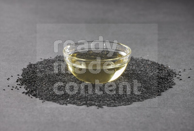 A glass bowl full of black seeds oil surrounded by the seeds on a black flooring