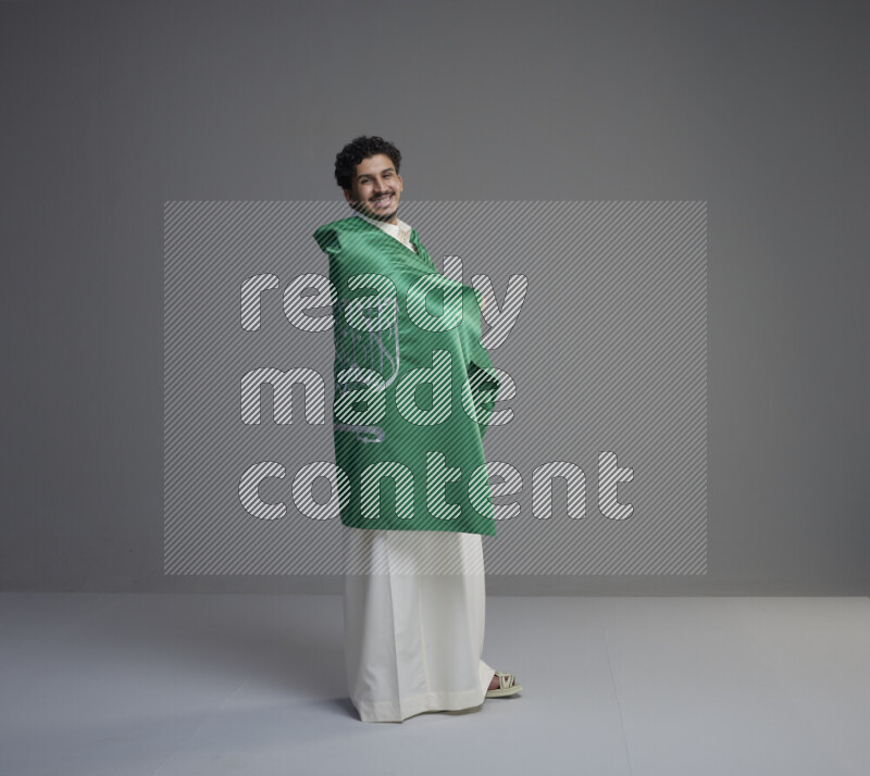 A Saudi man standing wearing thob wrapping big Saudi flag on gray background