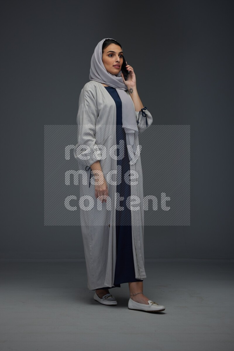 A Saudi woman wearing a light gray Abaya and head scarf standing and talking in the phone eye level on a grey background