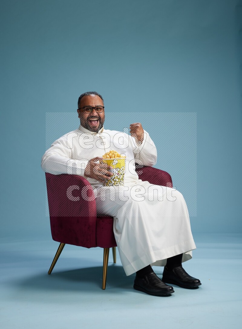 Saudi Man without shimag sitting on chair eating popcorn on blue background