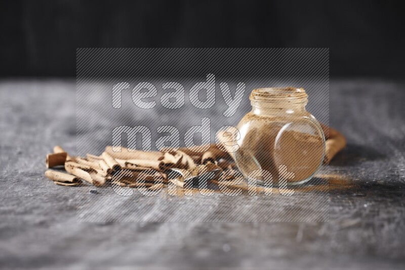 Herbal glass jar and a metal spoon full of cinnamon powder surrounded by cinnamon sticks on textured black background
