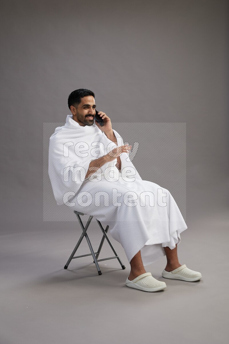 A man wearing Ehram sitting on chair talking on the phone on gray background