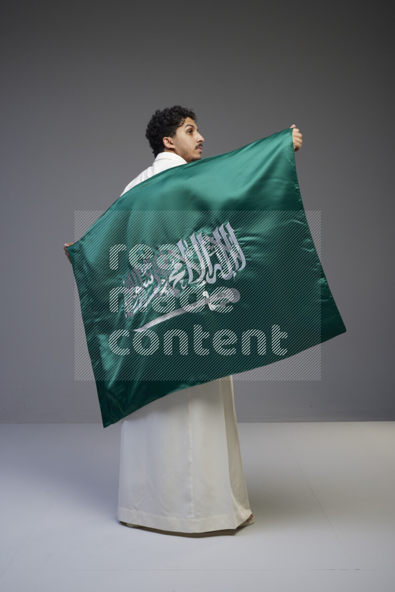A Saudi man standing wearing thob holding big Saudi flag on gray background
