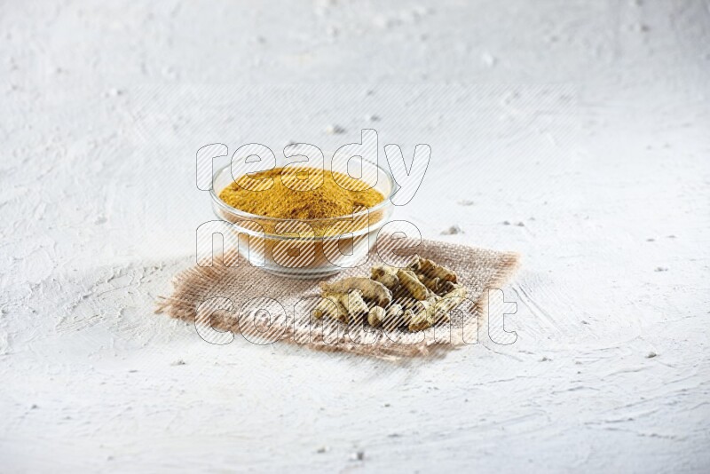 A glass bowl full of turmeric powder and dried turmeric whole finger on a piece of burlap on a textured white flooring