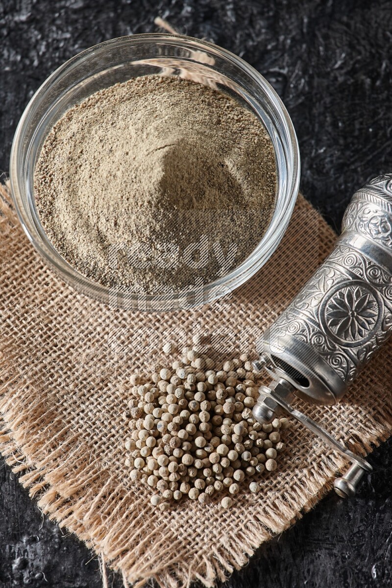 A glass bowl full of white pepper powder with white pepper beads on a burlap piece of fabric and a metal grinder on textured black flooring