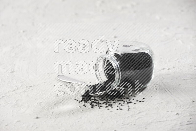 A glass spice jar and a metal spoon full of black seeds and the jar flipped and seeds spread on a textured white flooring