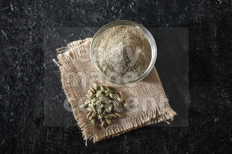 A glass bowl full of cardamom powder with cardamom seeds on a burlap piece on textured black flooring