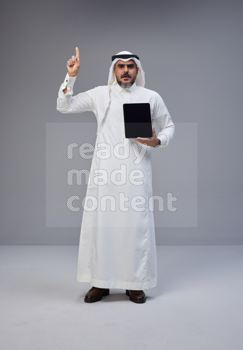 Saudi man Wearing Thob and white Shomag standing showing tablet to camera on Gray background