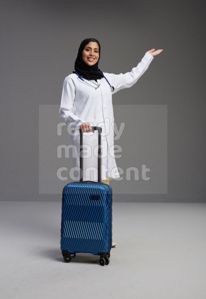 Saudi woman wearing lab coat with stethoscope standing holding Travel bag on Gray background