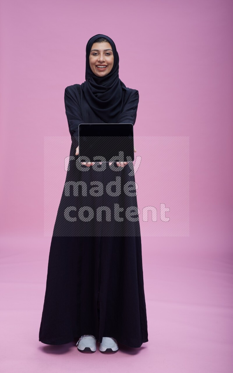 Saudi woman wearing Abaya standing showing tablet to camera on pink background