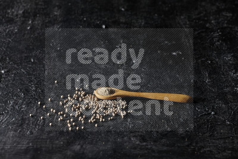 A wooden spoon full of white pepper powder with white pepper beads on textured black flooring