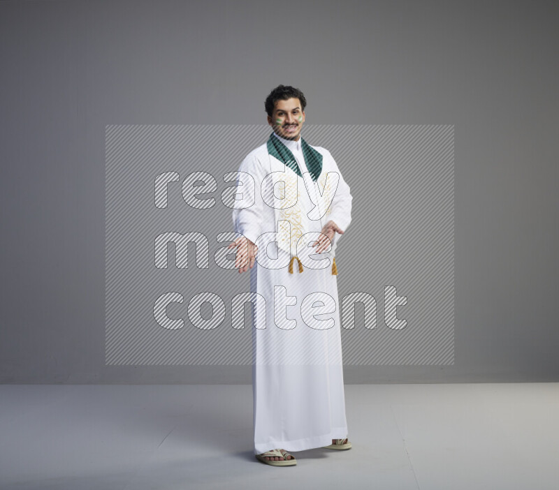 A Saudi man standing wearing thob and Saudi flag scarf with face painting on gray background