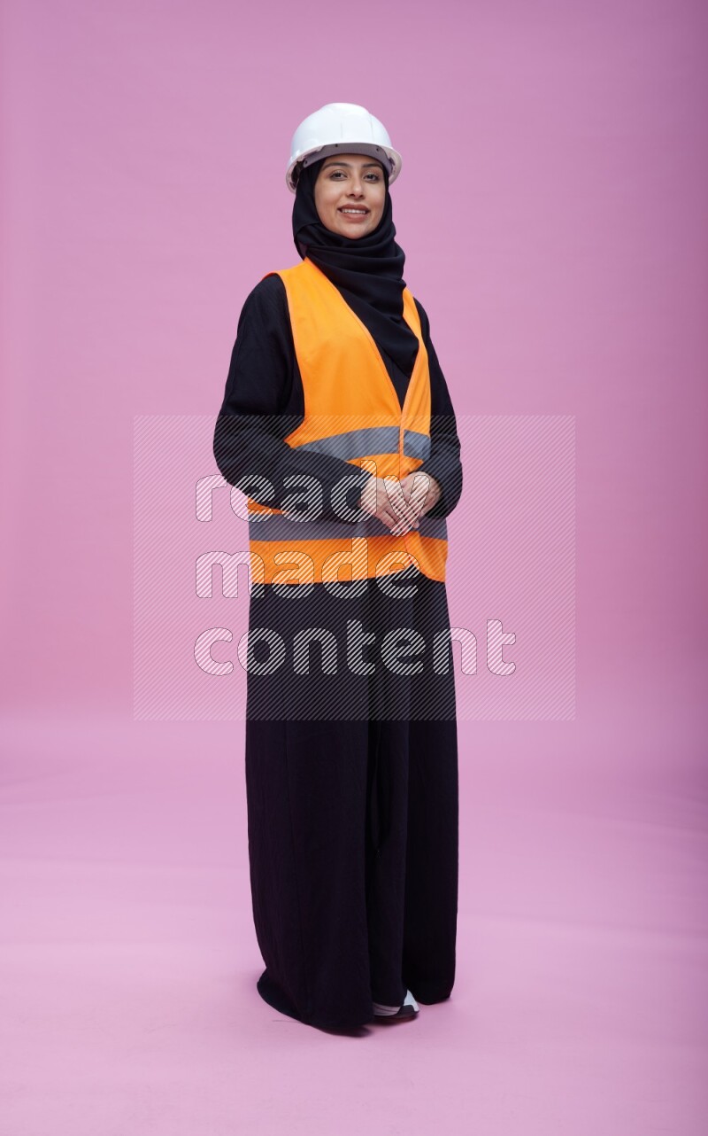 Saudi woman wearing Abaya with engineer vest and helmet standing interacting with the camera on pink background