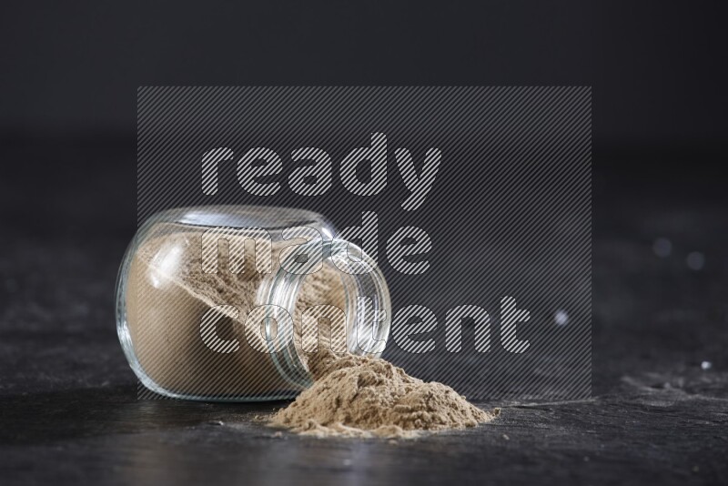 وعاء توابل زجاجي ممتلئ ببودرة الثوم مقلوب والبودرة متناثرة منه علي خلفية سوداء