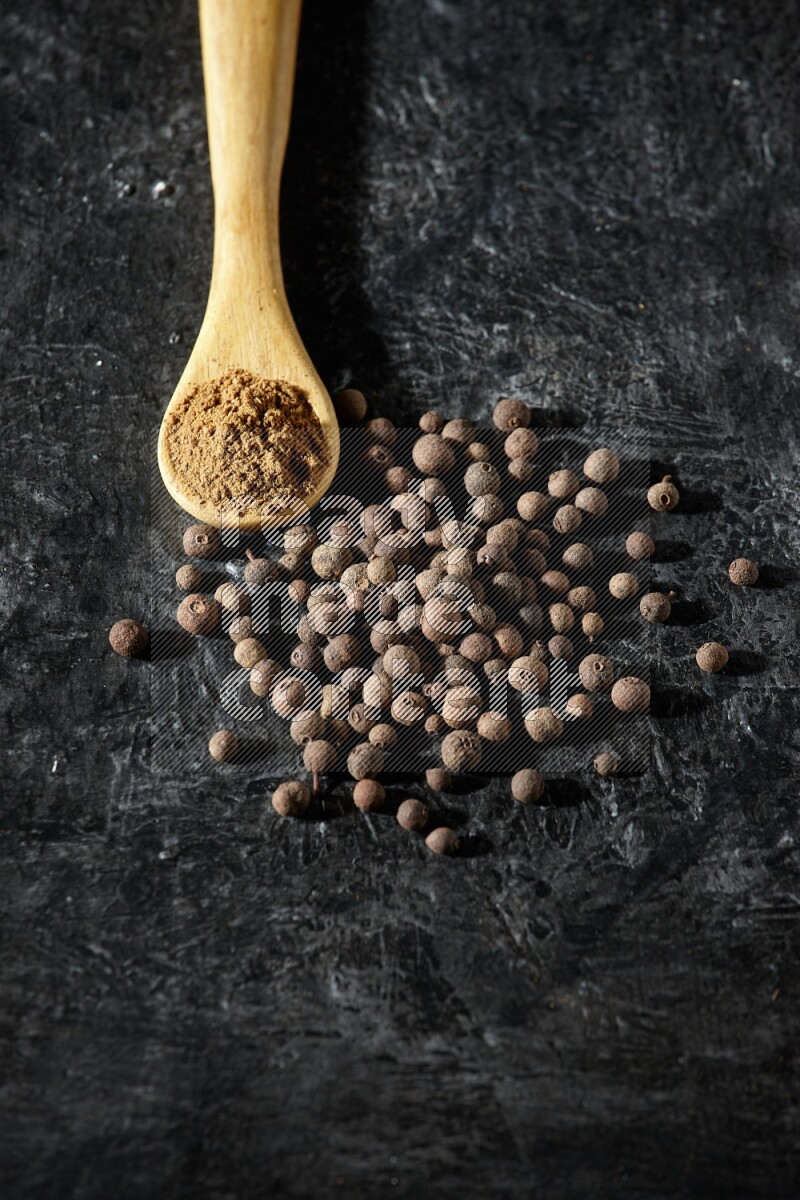 A wooden spoon full of allspice powder and whole balls spreaded on a textured black flooring