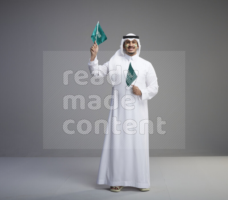 A Saudi man standing wearing thob and white shomag with face painting raising small saudi flag on gray background