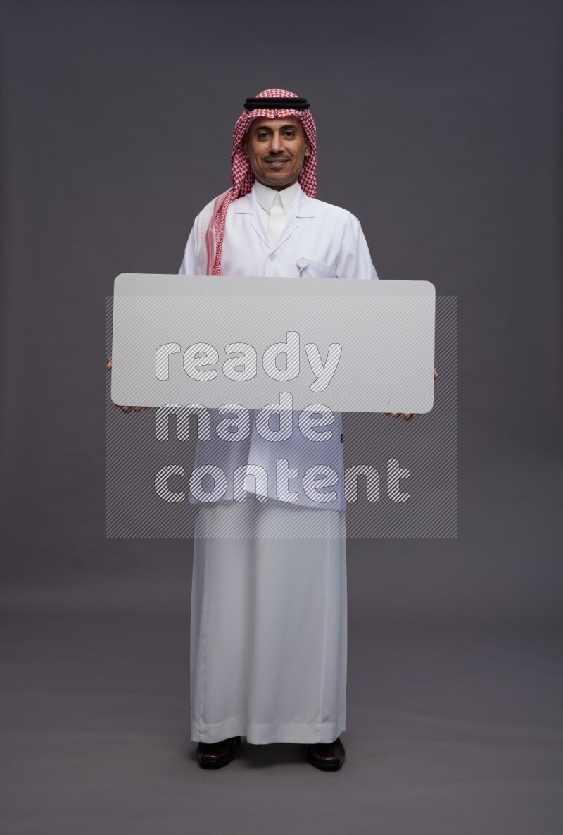Saudi man wearing thob with lab coat and shomag with pocket employee badge standing holding board on gray background