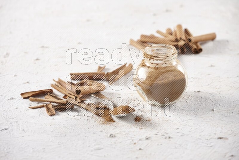 Herbal glass jar full cinnamon powder and a metal spoon surrounded by cinnamon sticks on a white background