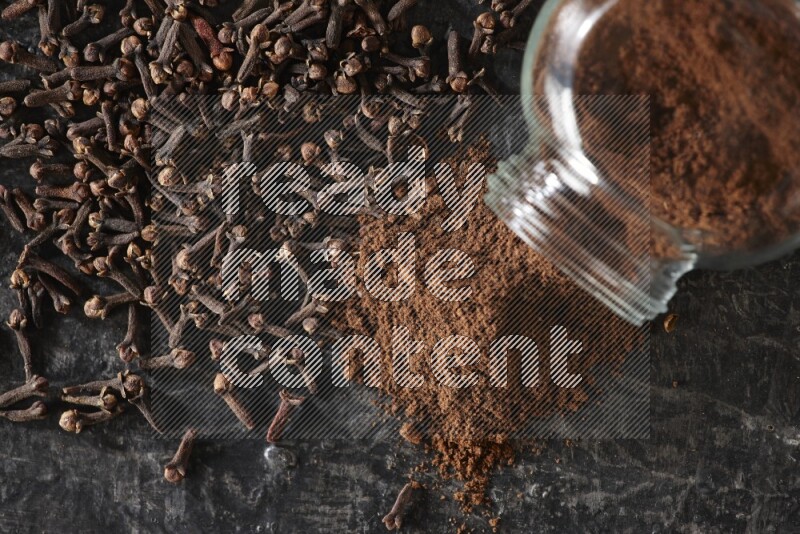 وعاء زجاجي للبهارات مقلوب ممتلئ ببودرة القرنفل والبودرة سقطت منه مع حبوب القرنفل المتناثرة على أرضية سوداء