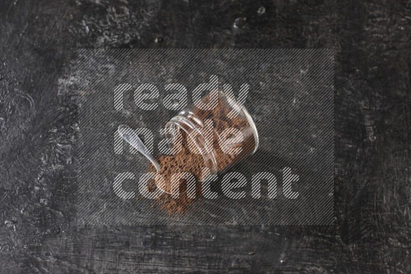 A flipped glass jar and metal spoon full of cloves powder with cloves spread on a textured black flooring