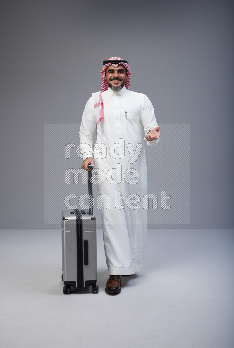 Saudi man wearing Thob and red Shomag standing holding Travel bag on Gray background