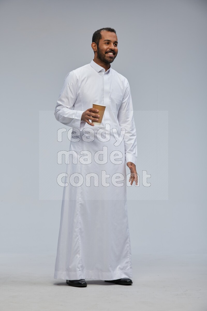 Saudi man wearing thob standing holding paper cup on gray background