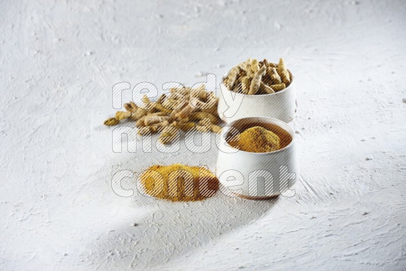 وعاءان فخاريان بيج ممتلئان ببودرة الكركم وأصابع الكركم المجففة، مع بودرة وأصابع الكركم بجانبهما على خلفية بيضاء