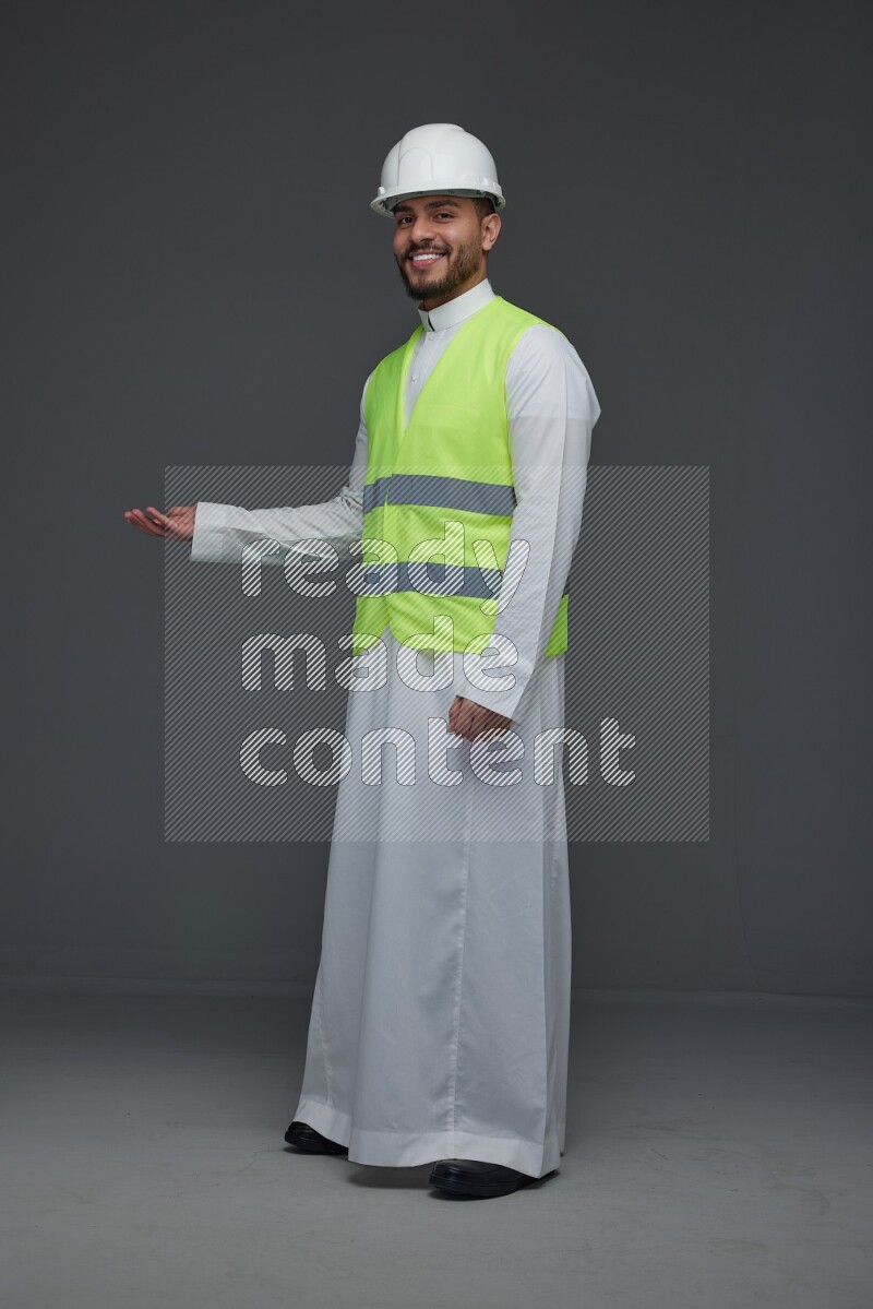 A Saudi man wearing Thobe with a yellow safety vest and white helmet standing and pointing different angles eye level on a gray background