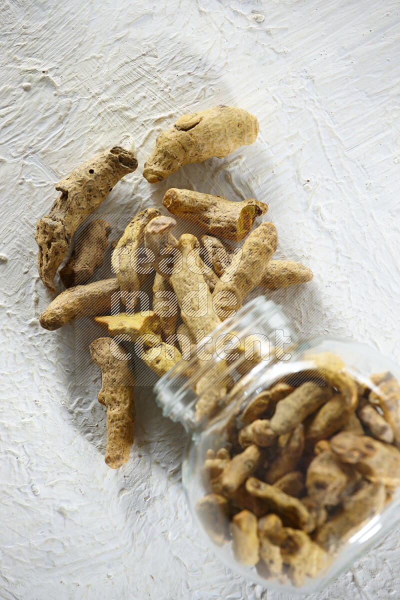A flipped glass spice jar full of dried turmeric fingers and fell out it on textured white flooring