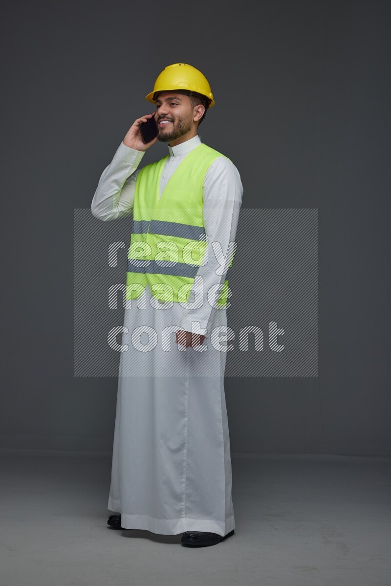 A Saudi man wearing Thobe with a yellow safety vest and white helmet standing and talking in the phone eye level on a gray background