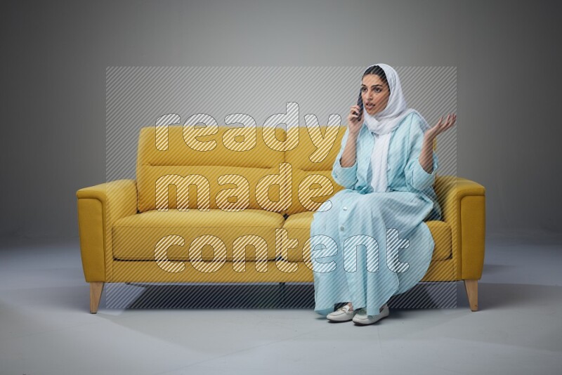 A Saudi woman wearing a light blue Abaya and a white head scarf sitting on a yellow sofa and talking in the phone while making different poses eye level on a grey background