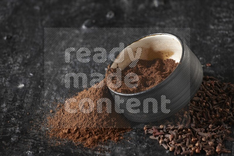 A black pottery bowl full of cloves powder spilled out of it with whole grains on a textured black flooring