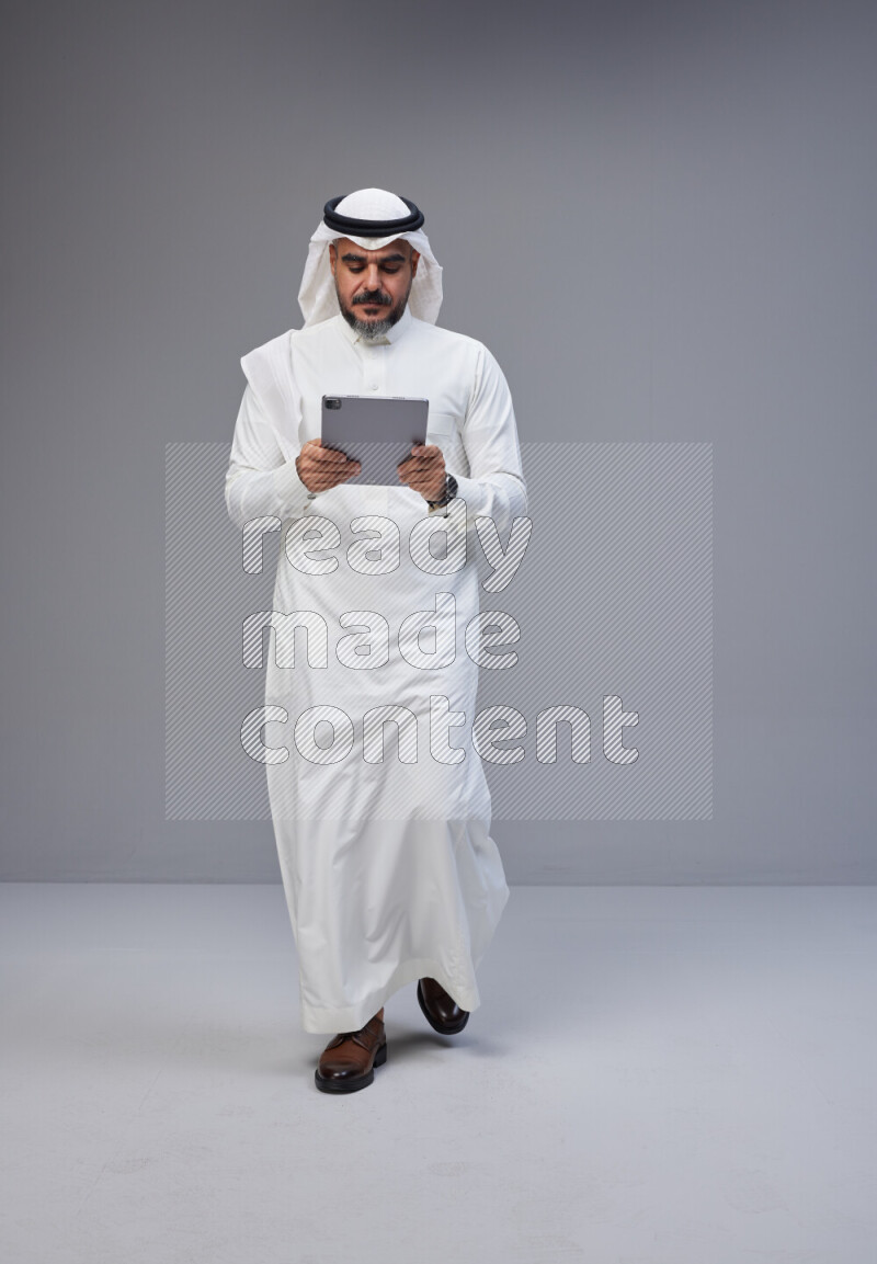 Saudi man Wearing Thob and white Shomag standing working on tablet on Gray background