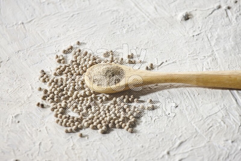 A wooden spoon full of white pepper powder and white pepper beads on textured white flooring