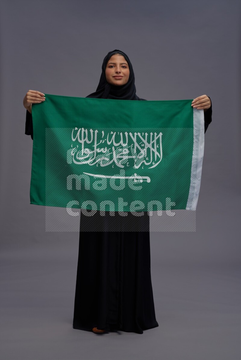 Saudi woman wearing Abaya standing holding Saudi flag on gray background