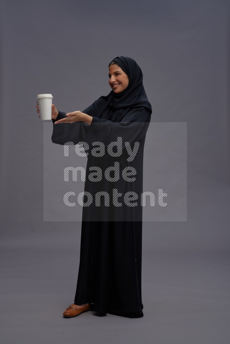 Saudi woman wearing Abaya standing holding paper cup on gray background