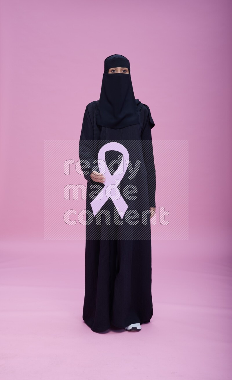 Saudi woman wearing abaya and niqab standing holding awareness ribbon on pink background