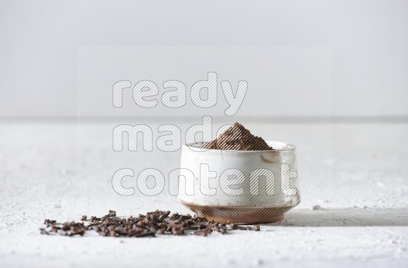 A beige ceramic bowl full of cloves powder and whole cloves on a white flooring