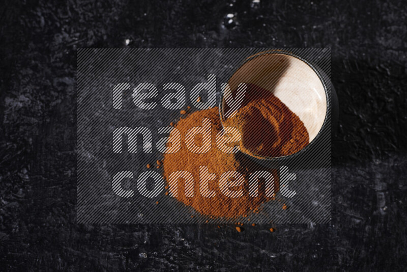 A black pottery bowl full of ground paprika powder with fallen powder from it on black background