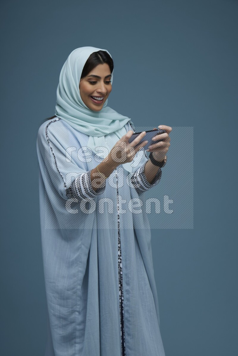 A Saudi woman Playing a Game on her phone on a blue background wearing a blue Abaya with hijab