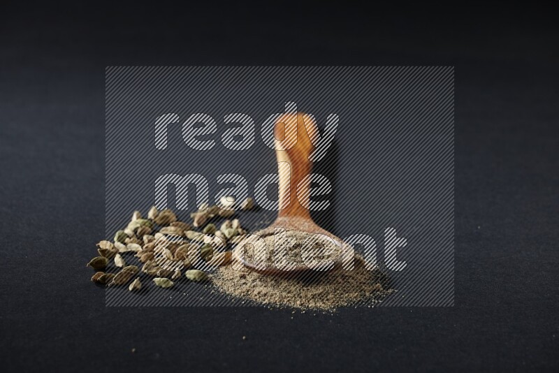 A wooden ladle full of cardamom powder and cardamom seeds beside it on black flooring