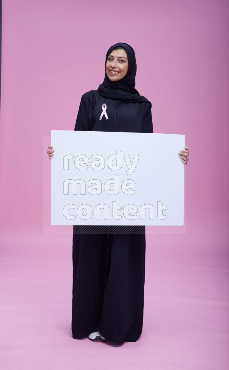 Saudi woman wearing pink ribbon on Abaya standing holding board on pink background