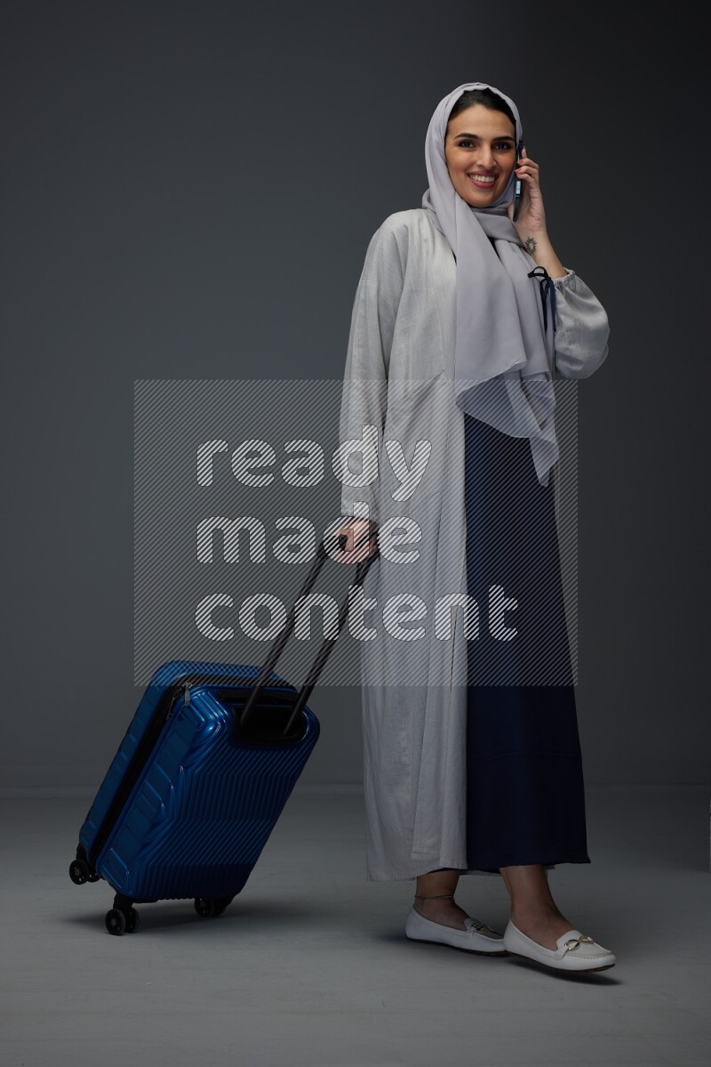 A Saudi woman wearing a light gray Abaya and head scarf standing and talking in the phone while holding shopping bags eye level on a grey background