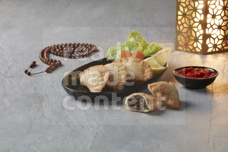 Three fried sambosas in an oval shaped black plate and a red sauce in a black round ramekin with a brown misbaha and a golden lantern on the side on a gray background