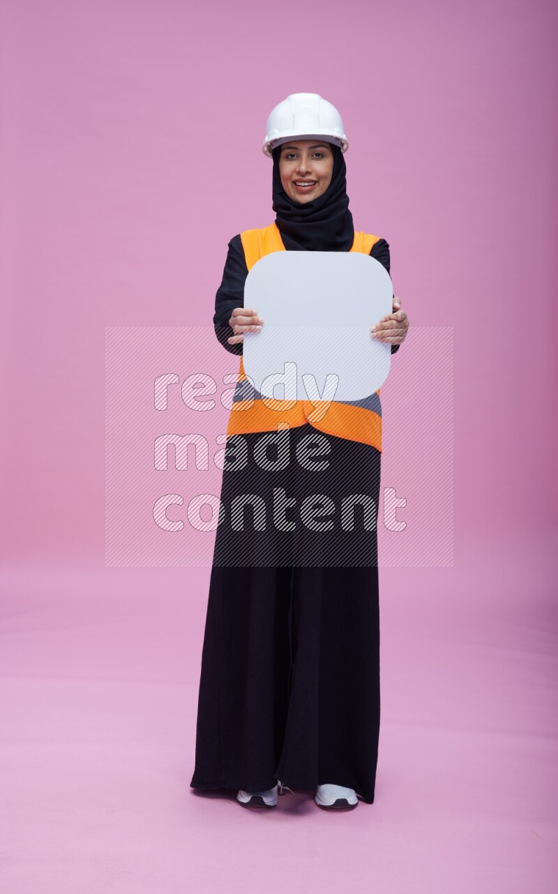 Saudi woman wearing Abaya with engineer vest and helmet standing holding social media sign on pink background