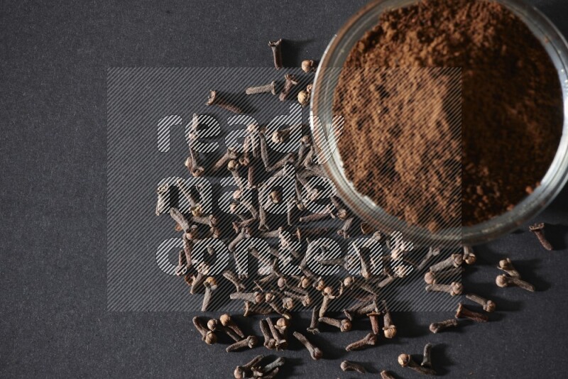 وعاء زجاجي ممتلئ ببودرة القرنفل مع حبوب القرنفل المتناثرة على أرضية سوداء