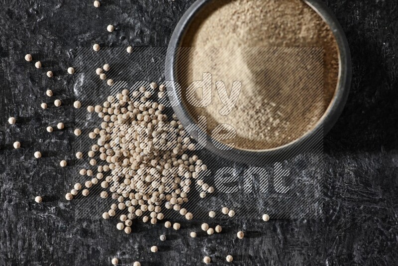 Black pottery bowl full of white pepper powder with white pepper beads on textured black flooring