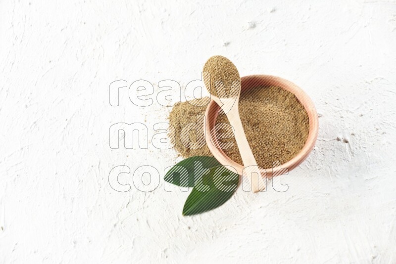 A wooden bowl and wooden spoon full of cumin powder on textured white flooring