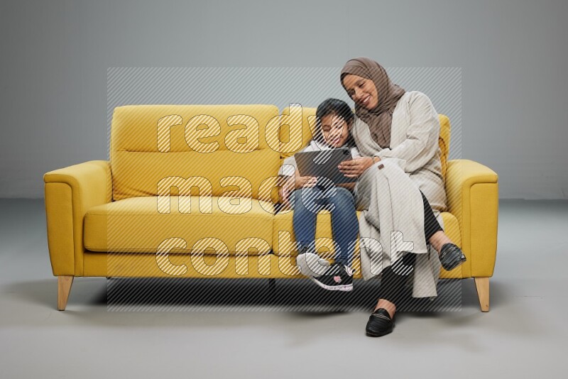 A girl with her mother on yellow sofa and watching on iPad on gray background