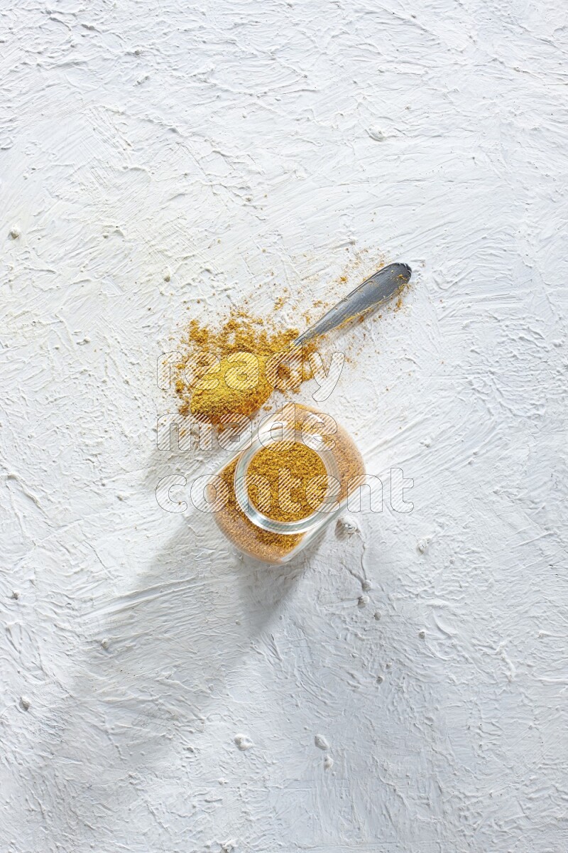 A glass spice jar and metal spoon full of turmeric powder on textured white flooring